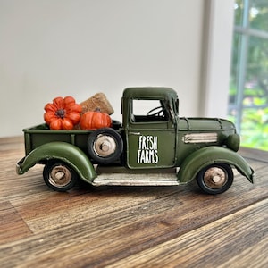 Pode incluir: Um camião de metal em miniatura de cor verde-azeitona com o texto FRESH FARMS impresso a branco na porta. A caixa do camião contém duas abóboras cor de laranja e um pequeno fardo texturado de cor bege. Tem rodas pretas e acabamentos prateados.