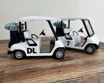 May include: Two white miniature golf carts with black accents and silver wheels. The front cart features the letters DL on the side, while the rear cart displays the number 7. Both vehicles carry a dark blue golf bag with a small green pine tree.