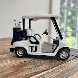 May include: A white miniature golf cart featuring black accents, a black steering wheel, and the letters TJ printed in black on the side. The vehicle includes a rear storage compartment holding a set of silver-colored golf clubs in a dark bag.
