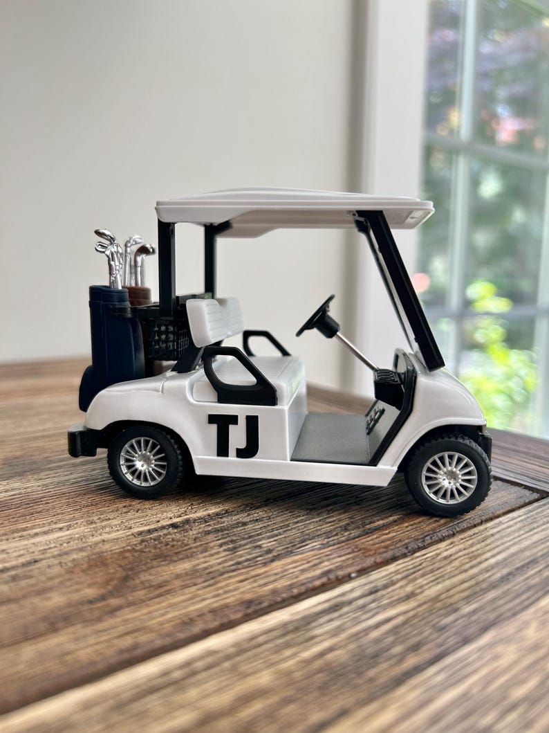 May include: A white miniature golf cart featuring black accents, a black steering wheel, and the letters TJ printed in black on the side. The vehicle includes a rear storage compartment holding a set of silver-colored golf clubs in a dark bag.