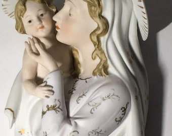 Peut inclure: Une figurine en porcelaine blanche de la Vierge Marie tenant un bébé Jésus. La figurine est décorée d'accents dorés et d'un motif floral. La Vierge Marie porte une robe blanche avec une auréole dorée.