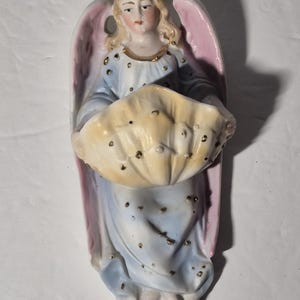 May include: A porcelain angel figurine with pink wings holding a yellow seashell. The angel is wearing a blue and white dress with gold accents.