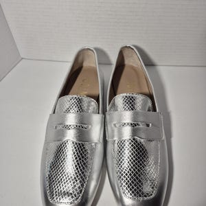 May include: A pair of silver leather loafers with a snakeskin pattern. The loafers have a rounded toe and a low heel.