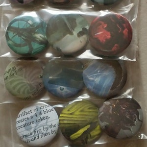 Peut inclure: Une collection de 15 boutons ronds et colorés avec différentes images et du texte. Les boutons sont disposés en rangées et emballés dans des sacs en plastique transparents. Le texte sur un bouton indique "artifact or creates a 4/4 blue creature token. struck first by the e ce, and then by"