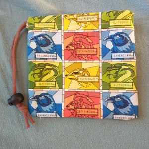 May include: A colorful fabric pouch with a drawstring closure. The pouch features a grid pattern with images of the Hogwarts house mascots: a lion, a badger, a snake, and an eagle. Each house is labeled with its name: Gryffindor, Hufflepuff, Slytherin, and Ravenclaw.