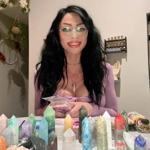 May include: A woman wearing glasses and a purple top sits at a table with a deck of tarot cards and a variety of colorful crystals. The crystals are arranged in a row on the table, with some of them standing upright and others lying flat.