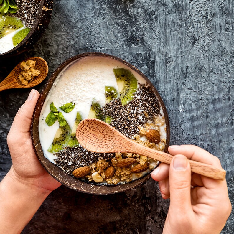May include: A close-up of a coconut bowl filled with a yogurt smoothie topped with kiwi, chia seeds, granola, and almonds. A wooden spoon is resting on the rim of the bowl.