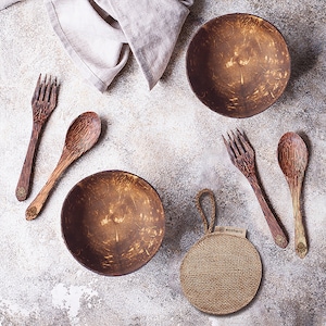 May include: A set of four brown coconut bowls and two wooden spoons and forks. The bowls are made from natural coconut shells and have a rustic, organic look. The spoons and forks are made from sustainable wood and have a natural finish. The set is displayed on a light gray background with a beige linen cloth and a brown burlap scrubby.