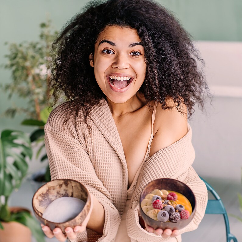May include: A woman in a beige robe holds two coconut bowls. One bowl contains a white smoothie, and the other bowl contains a fruit salad with mango, raspberries, blueberries, and blackberries.