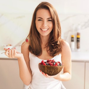 May include: A woman smiles while holding a coconut bowl filled with fresh raspberries and blueberries.