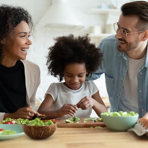 May include: A family of three preparing a meal together in a kitchen. A young girl is cutting cucumbers on a wooden cutting board. A woman is holding a bowl of salad. A man is smiling and looking at the girl. The family is using a coconut bowl for salad.