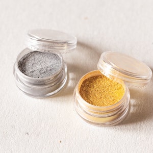 Könnte beinhalten: Zwei kleine durchsichtige Behälter mit Deckeln, einer gefüllt mit silbernem Glitter und der andere mit goldenem Glitter.
