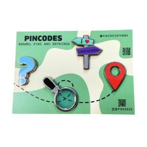 May include: A selection of enamel pins and keyrings on a green card. The pins feature a blue question mark, a magnifying glass, a red location marker, and a signpost with "Geocaching". The text "PINCODES" is at the top.