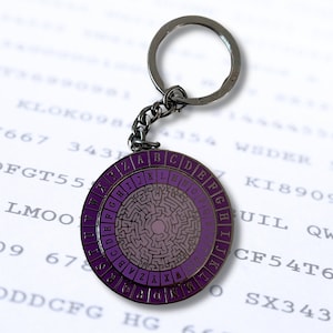 May include: A silver keychain with a purple and silver circular disc featuring a maze pattern and the letters of the alphabet around the edge. The keychain is attached to a silver key ring.