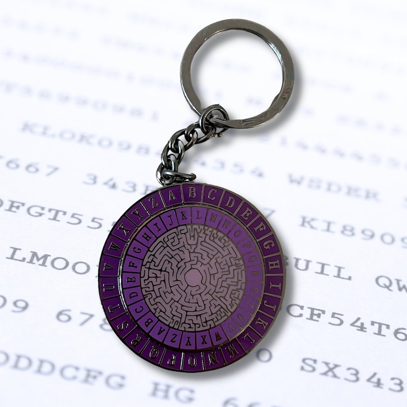 May include: A silver keychain with a purple and silver circular disc featuring a maze pattern and the letters of the alphabet around the edge. The keychain is attached to a silver key ring.