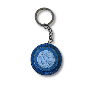 May include: A blue and white metal keychain with a circular design featuring a maze and the alphabet around the edge. The keychain has a silver metal ring.