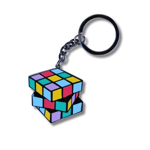 May include: A silver keychain with a colorful Rubik's Cube charm. The cube is made of metal and has a black outline. The colors of the cube are red, yellow, blue, green, pink, and purple.
