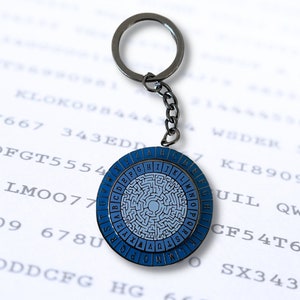 May include: A silver keychain with a blue and white circular cipher wheel. The wheel has a maze pattern and the letters of the alphabet are printed around the edge.
