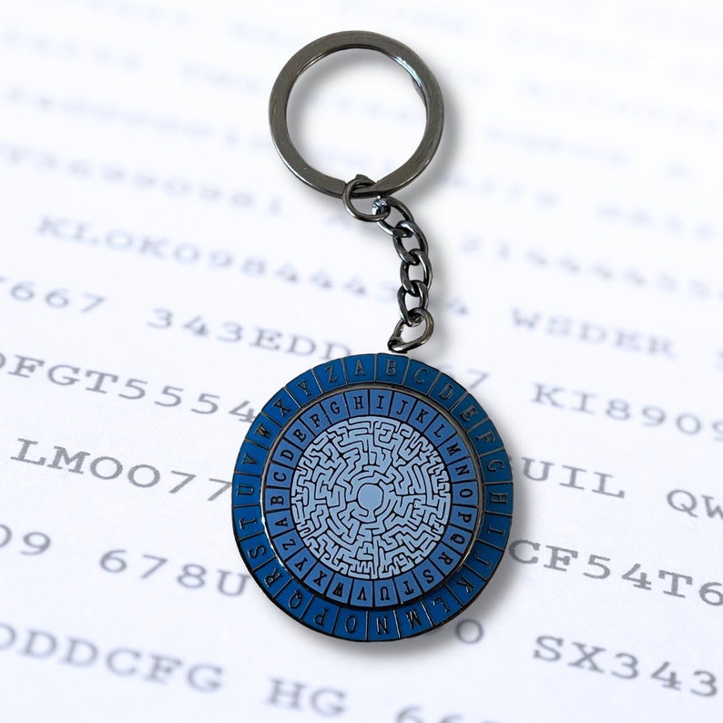 May include: A silver keychain with a blue and white circular cipher wheel. The wheel has a maze pattern and the letters of the alphabet are printed around the edge.