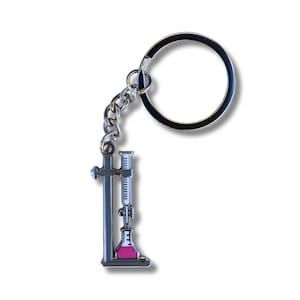May include: A silver keychain featuring a lab stand with a beaker and a burette. The beaker is filled with pink liquid.