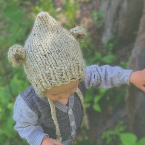 May include: A knitted beige hat with bear ears. The hat is worn by a child, who is wearing a gray sweater and a light blue long-sleeved shirt. The hat has ear flaps and ties. The background is blurred green foliage.