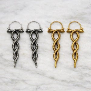 Può includere: Due paia di orecchini a cerchio con un design a doppio serpente. Un paio ha una finitura color argento, l'altro una finitura color oro. Ogni orecchino è composto da due serpenti intrecciati con squame testurizzate e un cerchio superiore.