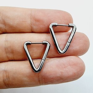 Puede incluir: Un par de pendientes de aro triangulares plateados. Los pendientes están hechos de metal y tienen un acabado liso.