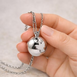Op de afbeelding: Een zilverkleurige hangerketting met een bolvormige bedel met een uitgesneden bladontwerp. De ketting wordt door een hand vastgehouden, waarbij de ketting en het gepolijste metaal van de hanger worden getoond. De achtergrond is een zachte, neutrale tint.