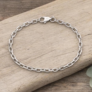 Può includere: Un braccialetto a catena color argento con un design a maglie ovali. Il braccialetto ha una chiusura a moschettone ed è contrassegnato con "925". Il gioiello è esposto su una superficie di legno.