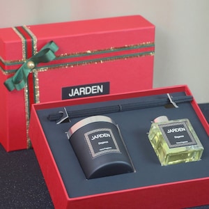 Op de afbeelding: Een rode cadeaudoos met een groene strik en een gouden lint, met daarin twee zwarte kaarsenpotten en een diffuser met het woord "JARDEN" op de doos en de potten. De potten hebben een zilveren deksel en de tekst "JARDEN Elegance Home Fragrance" op de voorkant.