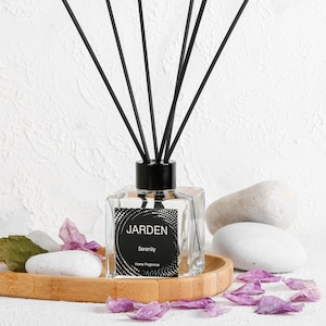 Puede incluir: Una botella de vidrio transparente con una tapa negra y palitos difusores negros. La botella tiene una etiqueta que dice "JARDEN Serenity Home Fragrance".