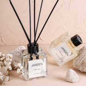 Può includere: Due bottiglie di vetro trasparente con tappi neri e bastoncini diffusori neri. Le bottiglie contengono un liquido giallo chiaro e hanno un'etichetta bianca con il testo "JARDEN Hommage Home Fragrance".