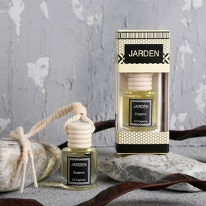 Puede incluir: Un ambientador de coche en una botella de vidrio transparente con una tapa de madera y una cuerda para colgar. La botella está etiquetada con "JARDEN Elegance Car Fragrance". El ambientador está empaquetado en una caja negra y dorada con la misma etiqueta.