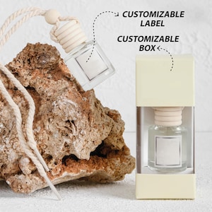 Może przedstawiać: Białe pudełko z przezroczystą szklaną butelką odświeżacza powietrza do samochodu w środku. Butelka ma białą etykietę i drewniany korek. Pudełko ma białą etykietę z napisem "CUSTOMIZABLE LABEL" i "CUSTOMIZABLE BOX".