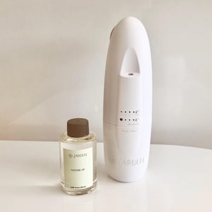 Op de afbeelding: Een witte diffuser met een zwart-wit label met de tekst "JARDIN" en een klein, doorzichtig glazen flesje met een zwarte dop en een label met de tekst "JARDIN NATURE UP".