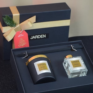 以下が含まれることがあります： 金色のリボンと「明けましておめでとうございます」と書かれた赤いギフトタグが付いた黒いギフトボックス。ボックスが開いており、金色の蓋が付いた黒いキャンドルと、金色の蓋が付いた透明なガラス製のディフューザーボトルが入っています。キャンドルとディフューザーの両方に「JARDEN」の文字が印刷されています。