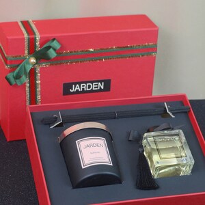 May include: A red gift box with a green bow and gold ribbon. The box contains a black candle with a silver lid and a clear glass bottle with a black tassel. The candle and bottle both have the word "JARDEN" on the label.