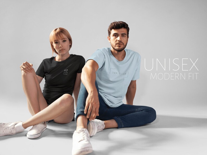 May include: Two people are wearing t-shirts with text on them. The person on the left is wearing a black t-shirt with the text "ADVENTURE" on it. The person on the right is wearing a light blue t-shirt with the text "TRAVELER" on it. The text "UNISEX MODERN FIT" is displayed in white text on a gray background.