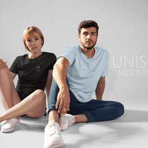 May include: Two people are wearing t-shirts with text on them. The person on the left is wearing a black t-shirt with the text "ADVENTURE" on it. The person on the right is wearing a light blue t-shirt with the text "TRAVELER" on it. The text "UNISEX MODERN FIT" is displayed in white on a gray background.