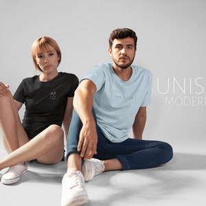 May include: Two people are wearing t-shirts with text on them. The person on the left is wearing a black t-shirt with the text "ADVENTURE" on it. The person on the right is wearing a light blue t-shirt with the text "TRAVELER" on it. The text "UNISEX MODERN FIT" is displayed in white on a gray background.