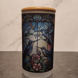 May include: A white ceramic jar with a wooden lid features a stained glass design of three blue hummingbirds and pink flowers.