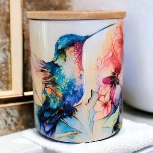 Puede incluir: Un tarro de cerámica blanco con tapa de bambú presenta una pintura de acuarela de un colibrí en vuelo con flores rosas y amarillas.