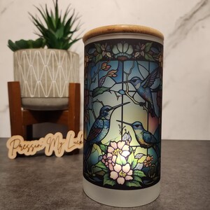 May include: A white ceramic candle holder with a bamboo lid. The candle holder features a stained glass design of three hummingbirds in flight with pink flowers.
