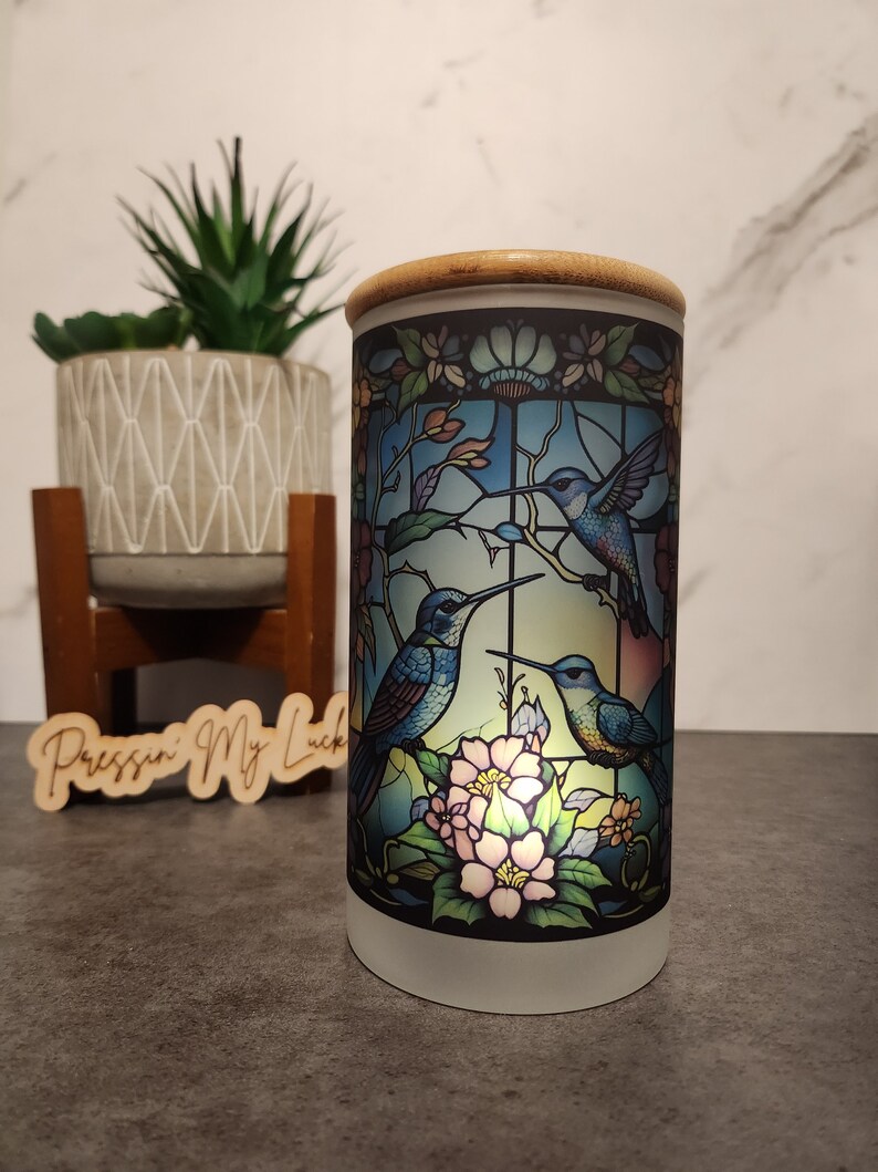 May include: A white ceramic candle holder with a bamboo lid. The candle holder features a stained glass design of three hummingbirds in flight with pink flowers.
