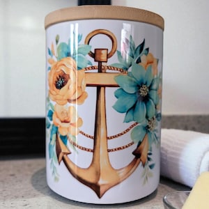 以下が含まれることがあります： 竹製の蓋が付いた白い円筒形の容器。オレンジとターコイズのフローラルエレメントに囲まれた金色のアンカーデザインで装飾されています。航海をテーマにした容器は、バスルームやキッチンでの使用に適しています。