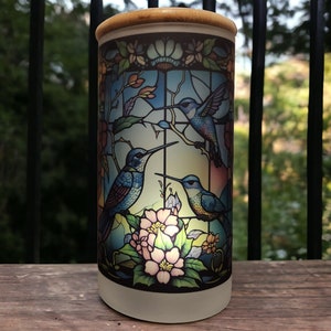 May include: A white ceramic candle holder with a stained glass design featuring three hummingbirds and pink flowers. The candle holder has a wooden top.