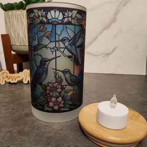 May include: A frosted glass candle holder with a stained glass design featuring three hummingbirds and flowers. The holder is paired with a small white LED tea light on a wooden base.