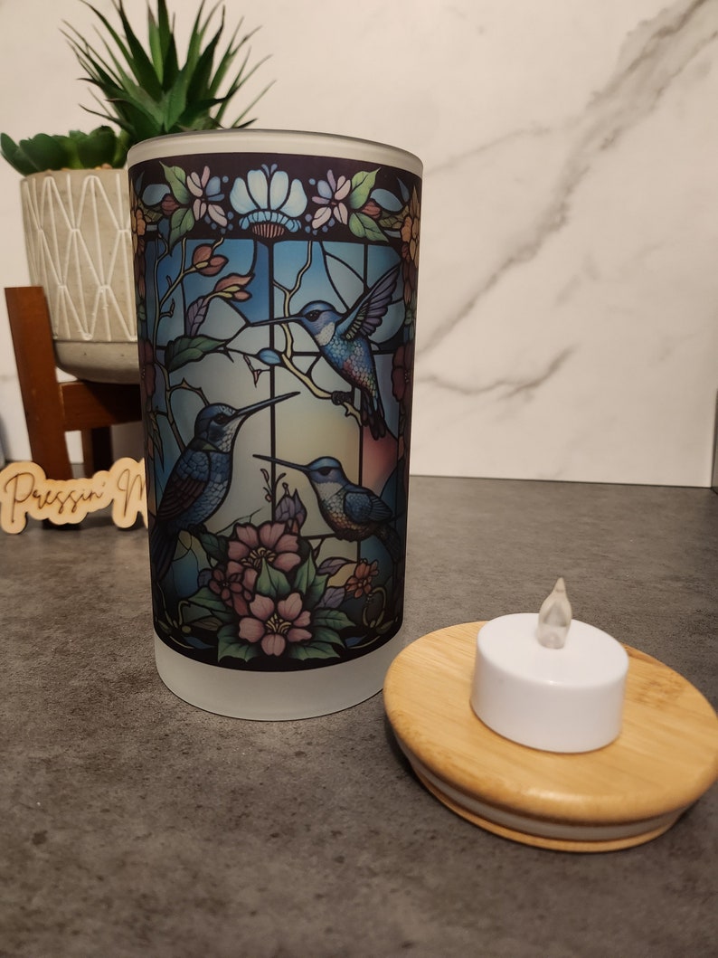May include: A frosted glass candle holder with a stained glass design featuring three hummingbirds and flowers. The holder is paired with a small white LED tea light on a wooden base.