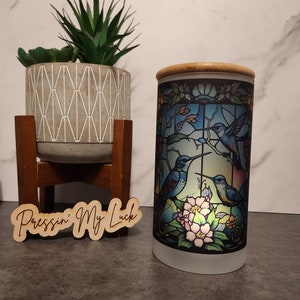 May include: A frosted glass jar with a bamboo lid features a stained glass design of three hummingbirds in flight with flowers. The jar is sitting on a gray surface with a wooden sign that reads "Pressin' My Luck".