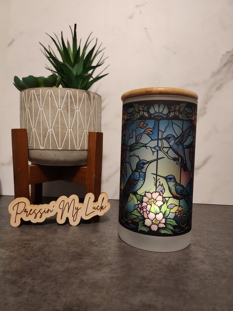 May include: A frosted glass jar with a bamboo lid features a stained glass design of three hummingbirds in flight with flowers. The jar is sitting on a gray surface with a wooden sign that reads "Pressin' My Luck".